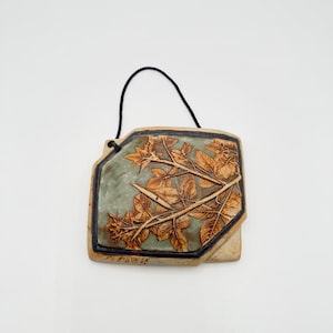 May include: A rectangular ceramic wall plaque with a sage green center and a raised, light brown relief of detailed leaves and branches. The piece is framed by a dark brown border and includes a black string for hanging on a wall.