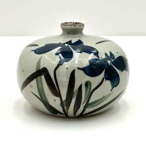 May include: A small, round ceramic vase with a white glaze and blue and brown floral designs. The vase has a narrow opening at the top.