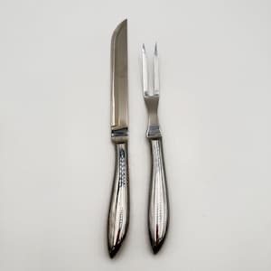 May include: A stainless steel carving knife and a two-pronged carving fork set. Both utensils feature smooth, polished silver-toned metal handles with a curved, ergonomic design and a sleek, reflective finish for serving roasted meats.