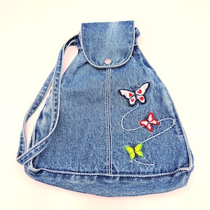 May include: A blue denim backpack with a drawstring closure and a flap secured by a silver snap. The backpack is decorated with embroidered butterflies in red, white, and yellow, connected by a white thread.