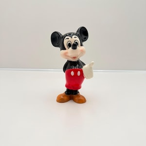 Pu&ograve; includere: Una piccola statuina vintage di Topolino. La figura &egrave; nera, bianca, rossa e marrone. Topolino fa il pollice in su. La statuina ha grandi orecchie nere, un viso bianco e pantaloncini rossi.
