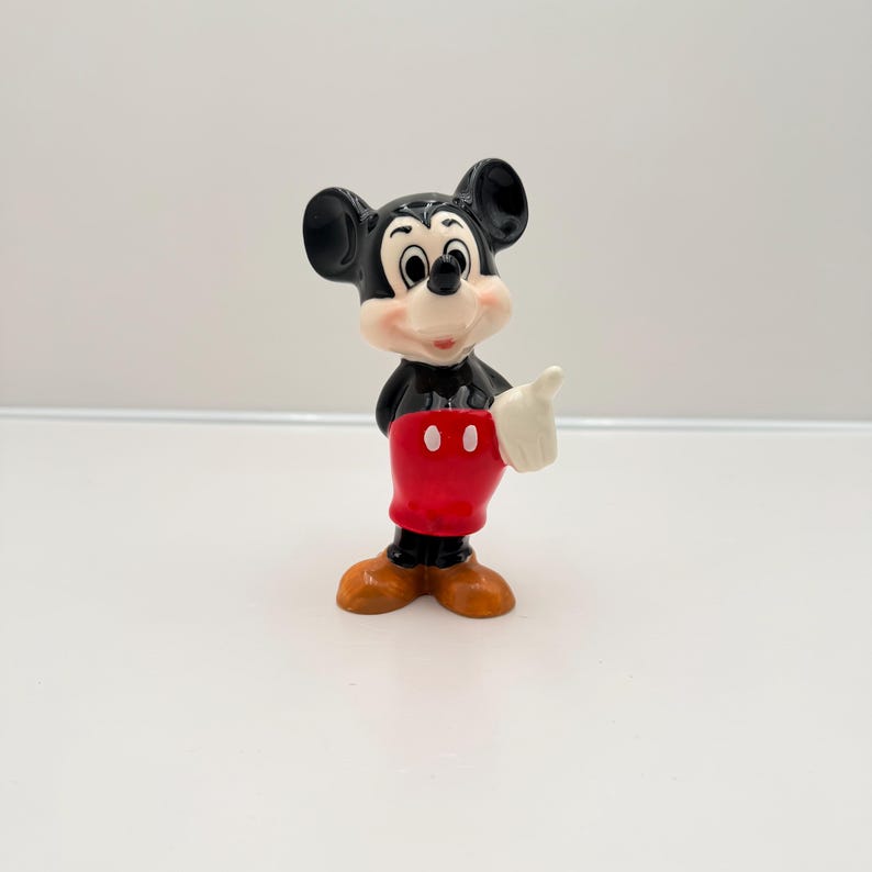 Pu&ograve; includere: Una piccola statuina vintage di Topolino. La figura &egrave; nera, bianca, rossa e marrone. Topolino fa il pollice in su. La statuina ha grandi orecchie nere, un viso bianco e pantaloncini rossi.