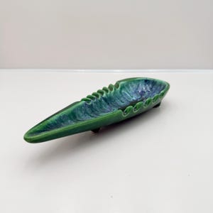 May include: A ceramic ashtray in a long, leaf-like shape. The ashtray is primarily green with blue and white accents, and has a textured, wavy edge. It has a glossy finish and is set on small feet.