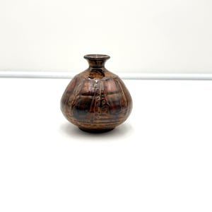 May include: A small, round ceramic vase with a narrow neck and a flared rim. The surface features a mottled glaze with blended tones of dark brown, copper, and black, creating a textured appearance with vertical drip-like patterns.