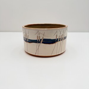 May include: A cylindrical ceramic bowl with a cream-colored glaze, featuring a horizontal dark blue band encircling the exterior. The surface is decorated with thin, hand-painted brown line drawings of cattail plants extending vertically across the bowl.