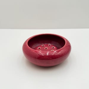 May include: A round, deep, glossy magenta ceramic flower frog bowl featuring a smooth, curved exterior and a flat interior base with seven evenly spaced circular holes designed for arranging stems in a floral display.