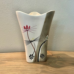 May include: A white ceramic vase with a geometric design and floral accents. The vase has a wide mouth and a narrow base. The floral design features pink and red flowers with black and green stems.