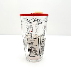 May include: A clear glass cocktail shaker with a red lid and a cream-colored top. The shaker features cocktail recipes and illustrations in black and white. The text includes "Whiskey Sour" and "Skoal!"