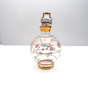 May include: A clear glass decanter with a round body and a matching stopper. The decanter is adorned with gold trim and a floral pattern in red, blue, and yellow. The stopper is also decorated with gold and floral accents.