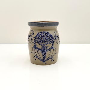 May include: A cylindrical stoneware crock with a tan body and a dark blue rim. The front features a hand-painted blue floral design with raised dot details and leaf accents. The vessel has a wide opening and a smooth, rounded base structure.