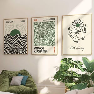 Puede incluir: Tres impresiones artísticas enmarcadas cuelgan de una pared blanca. Las impresiones presentan diseño Bauhaus, la obra de Yayoi Kusama y una figura de Keith Haring. Una silla puf verde con cojines se encuentra debajo, con plantas y un sofá blanco en primer plano.