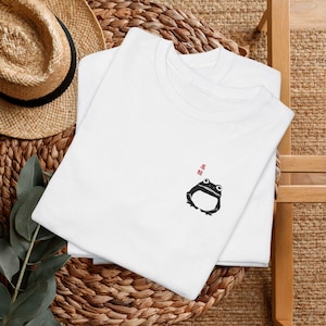 Puede incluir: Dos camisetas blancas apiladas, con un sombrero de paja en el fondo. La camiseta superior presenta un pequeño gráfico de rana negra y texto japonés rojo. La rana tiene ojos grandes y un cuerpo curvado. Las camisetas están sobre una superficie tejida.