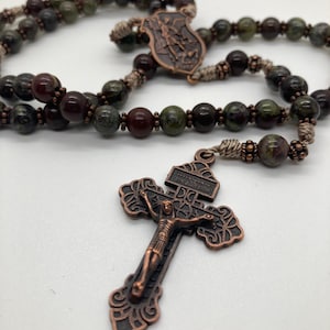 May include: A rosary with dark green and brown stone beads. The rosary features a bronze-colored crucifix with a detailed depiction of Jesus and a shield-shaped centerpiece. The beads are strung on a light brown cord.
