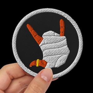 Puede incluir: Parche bordado circular con un diseño de gesto de mano de rock and roll. Presenta dedos naranjas, una mano envuelta en blanco y un fondo negro, todo dentro de un borde gris. También se ve un pequeño anillo dorado.