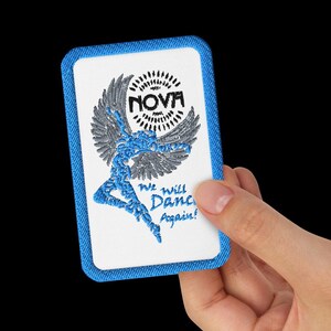 Peut inclure: Un écusson rectangulaire avec une bordure en jean bleu et un centre blanc. Le centre présente une danseuse stylisée en bleu, des ailes grises et le mot "NOVA" au-dessus. Le texte "We will Dance Again!" est en dessous.