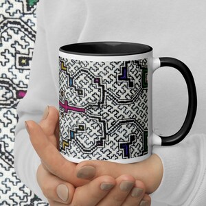 May include: A white ceramic mug with a black interior and handle, featuring a complex, repeating geometric pattern in black lines with small accents of pink, blue, yellow, and green. The design is printed on the exterior surface of the mug.