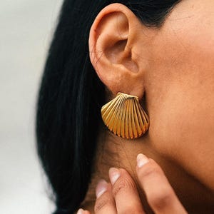 May include: A gold-toned earring shaped like a scallop shell with vertical ridges and a matching gold-toned ring featuring a twisted, rope-like texture. Both pieces of jewellery have a polished, metallic finish and a distinct, structured design.