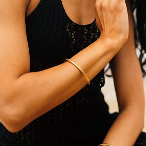 May include: A smooth, polished gold-tone metal bangle bracelet worn on the forearm, paired with a matching gold-tone ring featuring a series of small, rounded, connected spheres, both displayed against a black textured sleeveless garment.