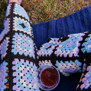 Op de afbeelding: Een persoon draagt een blauwe en paarse gehaakte broek met een glas rode sap op een blauwe deken.