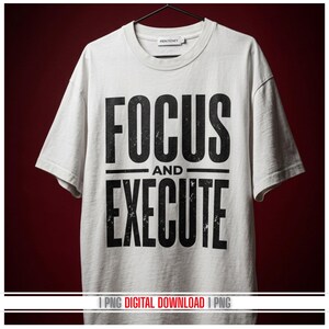May include: A white t-shirt with the words "FOCUS AND EXECUTE" printed in large, distressed black letters. The shirt is hanging on a black hanger against a dark red background. The text is centered on the shirt.