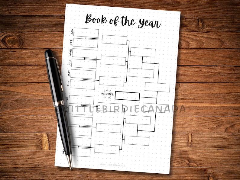May include: A black pen rests on a white paper with a bracket-style chart labeled "Book of the year." The chart has boxes for entries and a "Winner" section. The paper is dotted and sits on a wooden surface.