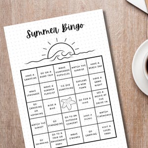 May include: A black and white printable summer bingo card with activities like making lemonade, going on a road trip, and watching fireworks. The card has a sun and waves graphic at the top.
