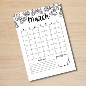 May include: A white March calendar page with a butterfly border. The calendar grid is blank, with days of the week labeled across the top. Includes space for monthly goals and notes. The page is on a light wood surface.