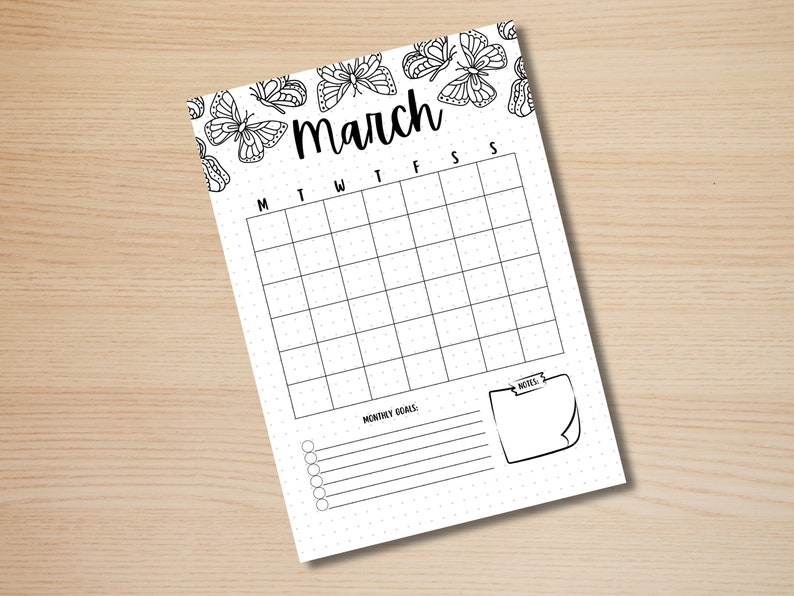 May include: A white March calendar page with a butterfly border. The calendar grid is blank, with days of the week labeled across the top. Includes space for monthly goals and notes. The page is on a light wood surface.