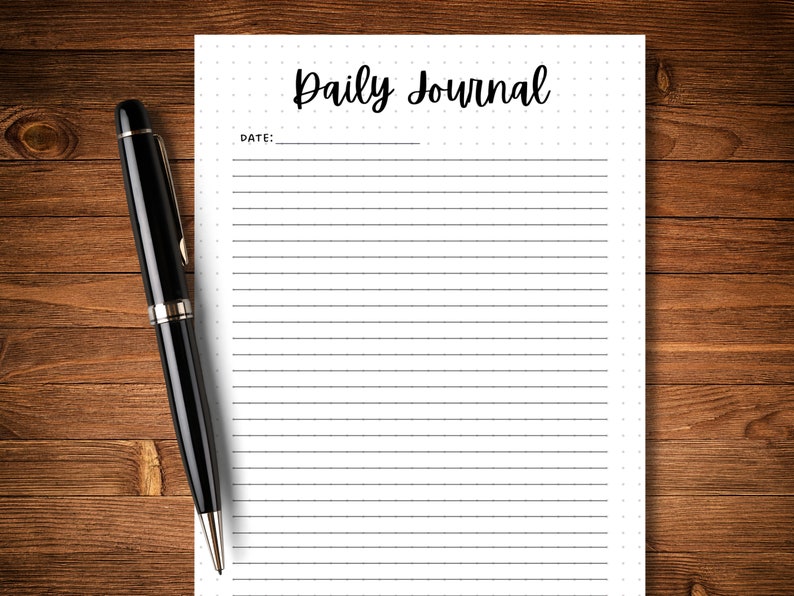 May include: A blank white lined journal with the text "Daily Journal" at the top and the word "DATE" followed by a blank line below. A black pen is lying on a wooden surface next to the journal.