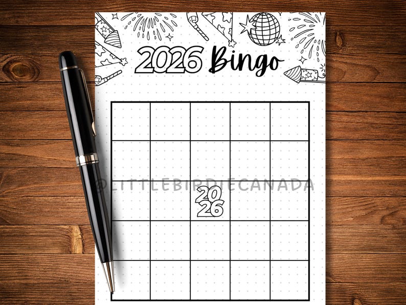 May include: A black pen lies next to a white bingo card. The card has a grid and the text "2026 Bingo." The top of the card features illustrations of fireworks and party hats. The background is a wooden surface.
