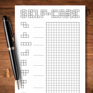May include: A black pen rests on a white self-care journal with a grid and the words "SELF-CARE" at the top. The journal has a series of shapes and lines for tracking habits. The background is a wooden surface.