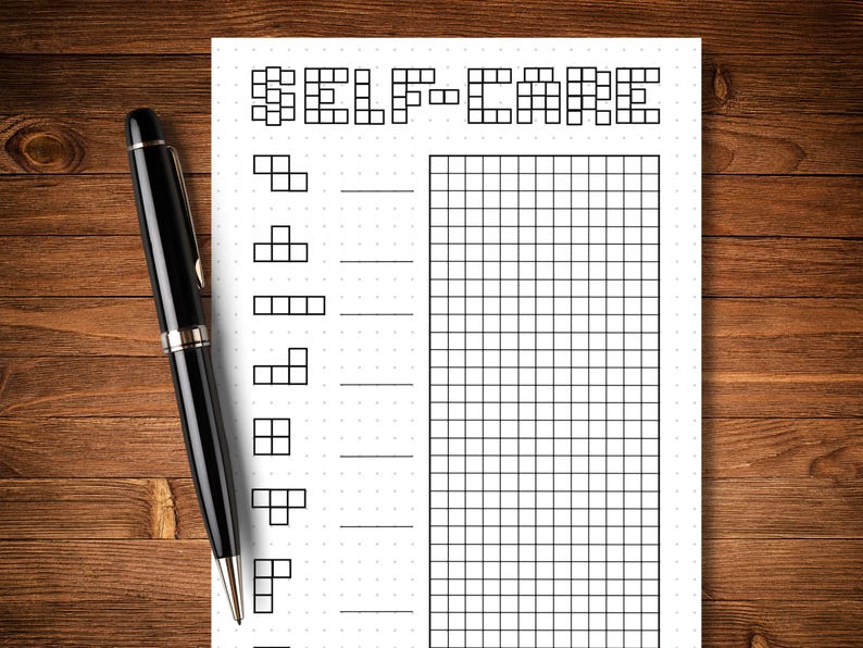 May include: A black pen rests on a white self-care journal with a grid and the words "SELF-CARE" at the top. The journal has a series of shapes and lines for tracking habits. The background is a wooden surface.