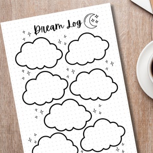 May include: A white Dream Log journal page with cloud-shaped sections for recording dreams. The page has a dotted background and is decorated with stars and a crescent moon with stars. A cup of coffee sits on a saucer to the right.