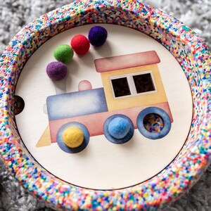 Könnte beinhalten: Ein rundes, farbenfrohes Tablett aus Kunststoff mit einem Zugmotiv in der Mitte. Der Zug ist blau, rot und gelb auf weißem Hintergrund. Um den Zug herum befinden sich kleine, bunte Kugeln.