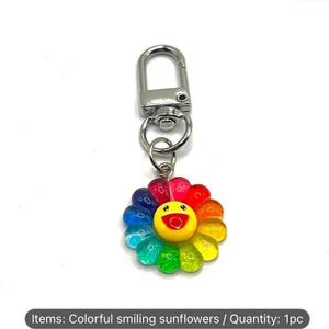 May include: A colorful keychain featuring a smiling sunflower design. The flower has vibrant petals in shades of red, orange, yellow, green, blue, and purple. It has a silver-colored metal clasp for attachment.