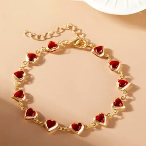 Op de afbeelding: Goudkleurige armband met hartvormige rode accenten. De armband heeft een delicaat kettingontwerp met meerdere hartvormige bedels. De sluiting en ketting zijn goudkleurig en de harten zijn felrood. Een romantisch en elegant sieraad.
