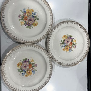 Puede incluir: Tres platos de cerámica blanca vintage con un diseño floral. Cada plato presenta un ramo de flores coloridas en el centro, incluyendo flores moradas, amarillas, naranjas y azules. Los bordes están adornados con un patrón de filigrana dorada, añadiendo un toque de elegancia.