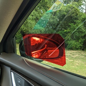 May include: A red tinted window film covers the side view mirror of a car. The film is shaped like a hexagon and is applied to the outside of the mirror. The mirror reflects a view of trees and a road.