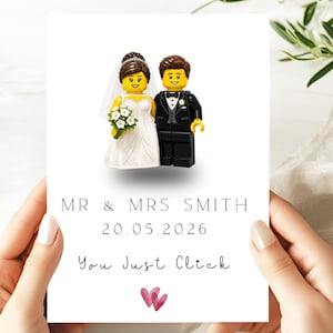 May include: A white wedding card featuring a Lego bride and groom figurine. The text reads MR & MRS SMITH, 20.05.2026, and You Just Click, with two small pink hearts at the bottom. The bride wears a white dress and veil, and the groom wears a black suit.
