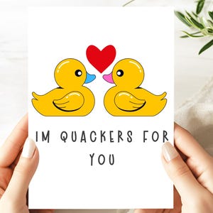 May include: A greeting card held by hands, featuring two yellow cartoon ducks with a red heart between them. The text "I'M QUACKERS FOR YOU" is printed below the ducks. The card is on a white background.