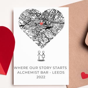 May include: A white card with a black and white heart-shaped map, a red heart, and the text "WHERE OUR STORY STARTS ALCHEMIST BAR - LEEDS 2022". A brown envelope and a red paper heart are also visible.
