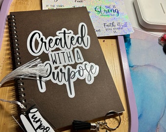 May include: A brown notebook with a white sticker that says "Created with a Purpose". There are several other stickers with inspirational quotes on a white background. There is also a white bookmark with black text that says "Purposefully Created" and a clear keychain with the text "Created Purpose".