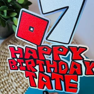 May include: A red and white cake topper with the number 7, a red square with a smaller square inside, and the words "Happy Birthday Tate".