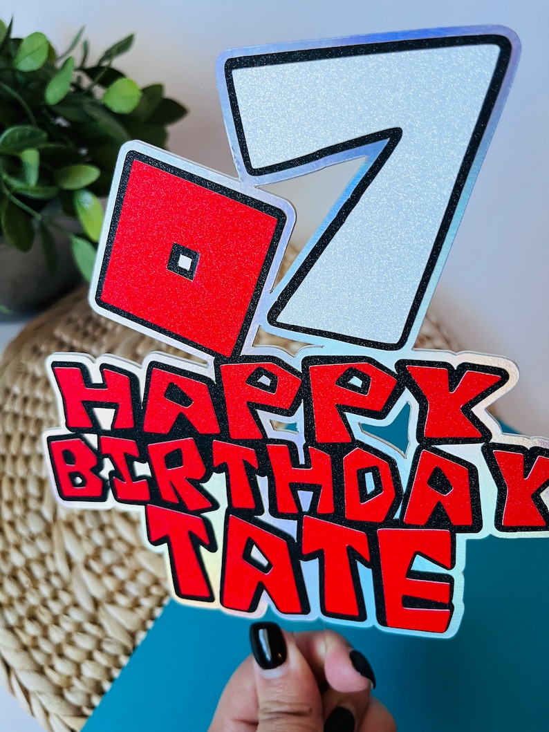 May include: A red and white cake topper with the number 7, a red square with a smaller square inside, and the words "Happy Birthday Tate".