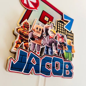 May include: A Roblox themed cake topper with a red and white Roblox logo, a blue number 7, and the name Jacob in blue and white letters. The topper also features several Roblox characters, including a robot, a construction worker, and a knight.