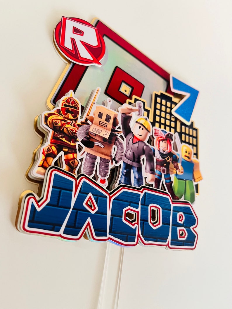 May include: A Roblox themed cake topper with a red and white Roblox logo, a blue number 7, and the name Jacob in blue and white letters. The topper also features several Roblox characters, including a robot, a construction worker, and a knight.