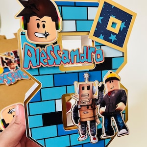 May include: A blue and gold Roblox themed cake topper with a brick pattern, a Roblox character with brown hair, a robot, and a Roblox character wearing a black jacket. The cake topper also includes the name Alessandro.