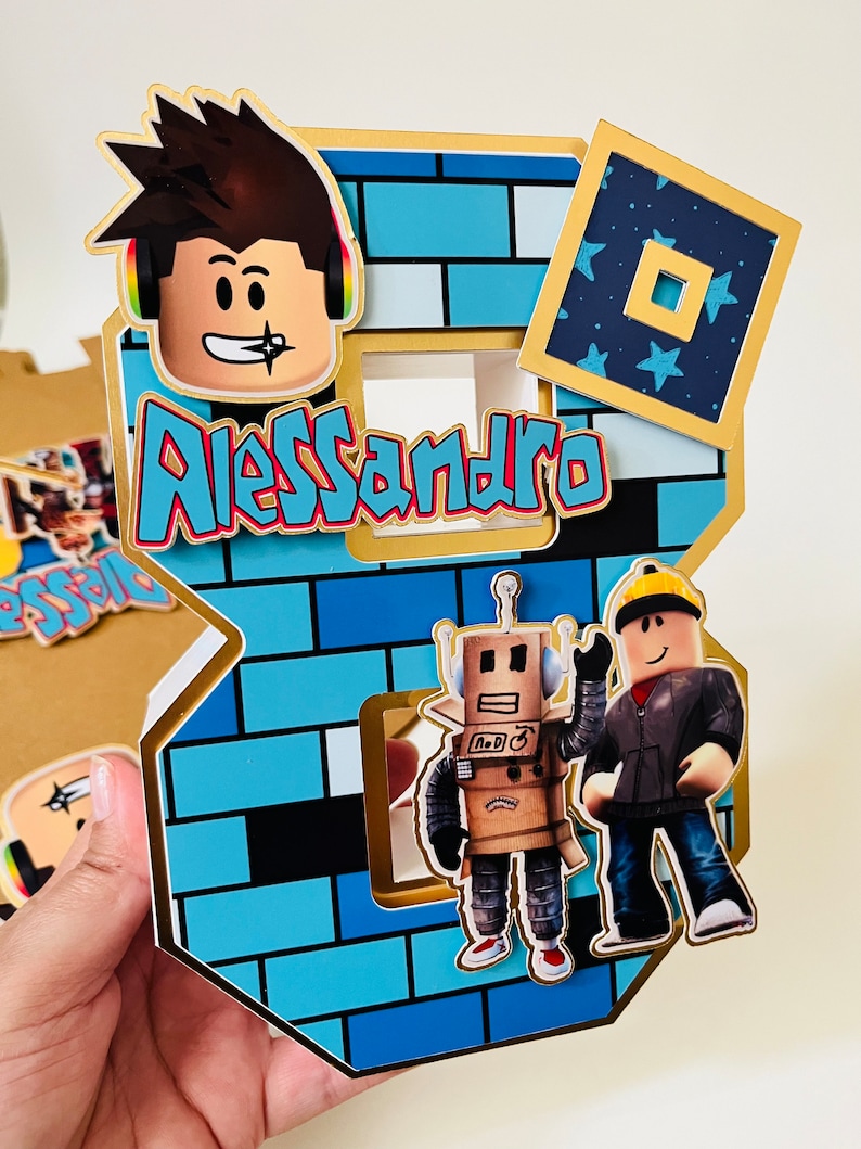 May include: A blue and gold Roblox themed cake topper with a brick pattern, a Roblox character with brown hair, a robot, and a Roblox character wearing a black jacket. The cake topper also includes the name Alessandro.