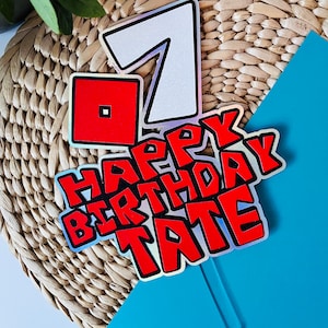 May include: A cake topper with a holographic finish featuring the number 7, a red Roblox logo, and the text "Happy Birthday Tate" in red and black.