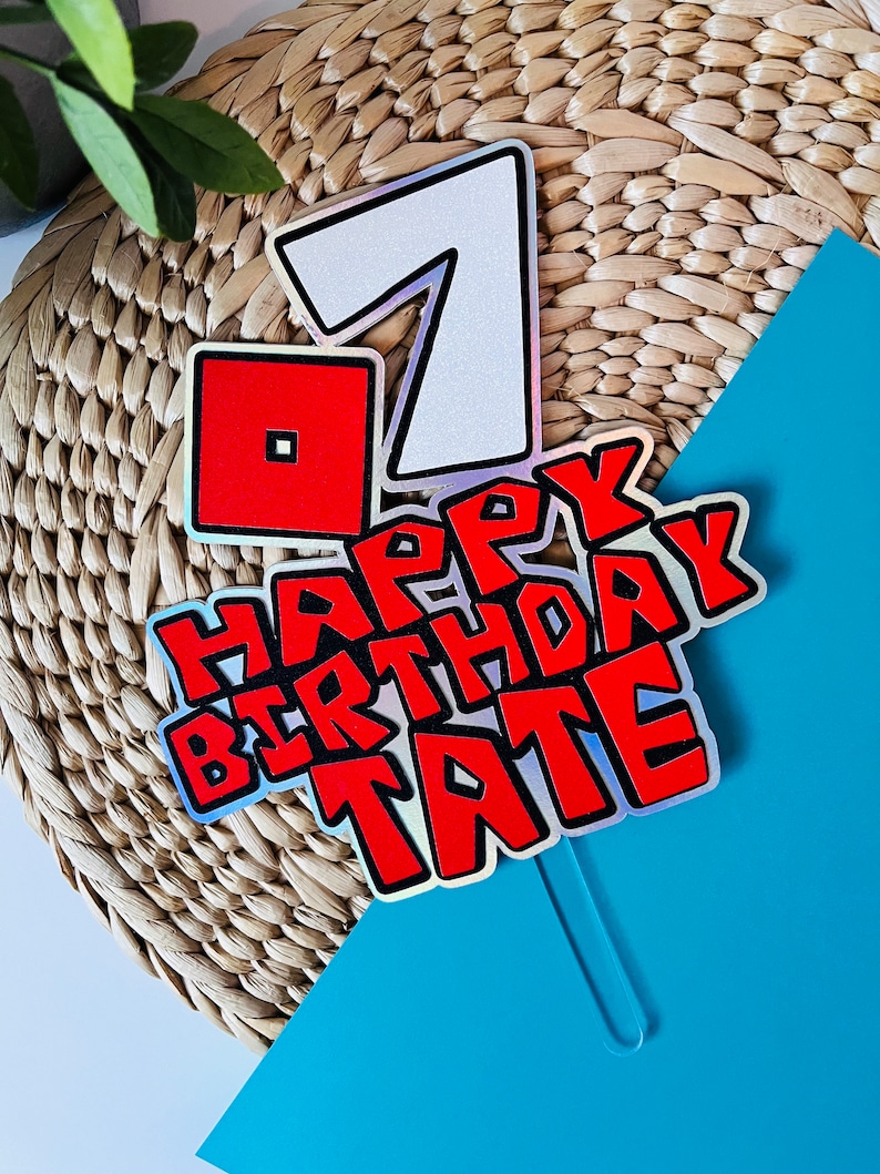 May include: A cake topper with a holographic finish featuring the number 7, a red Roblox logo, and the text "Happy Birthday Tate" in red and black.